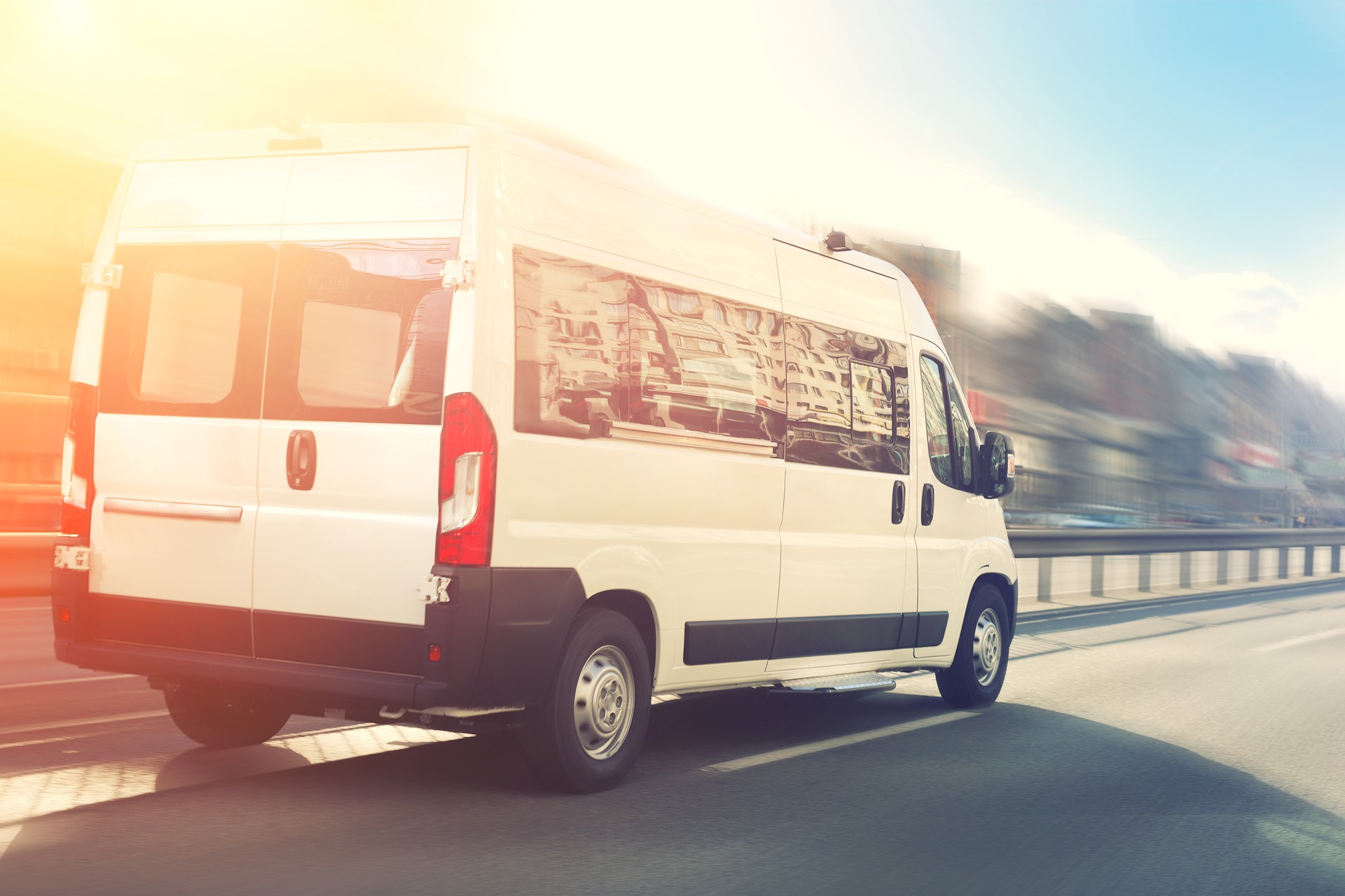 Unrecognizable small passenger van hurry up on highway at city street traffic with urban cityscape and sunset sky on background. Charter or shuttle bus  van hurry up on road with motion blur effect